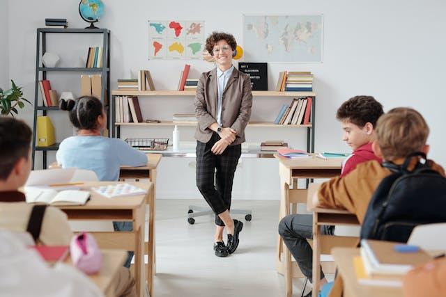 Groep kinderen zit aan tafels, voor hen een leraar.